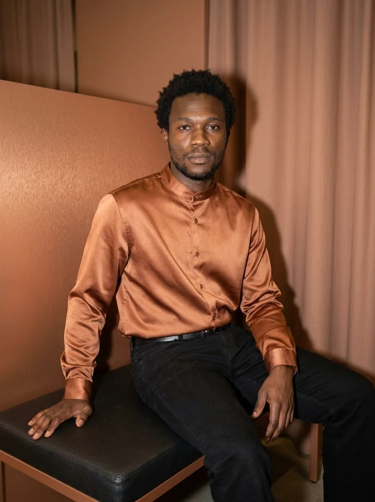 A fictional adult in a copper satin shirt and black jeans sits on a padded black bench, one hand on the seat. The wall and curtain behind are copper-toned, and bright frontal light casts a shadow on the curtain. AI-generated example.
